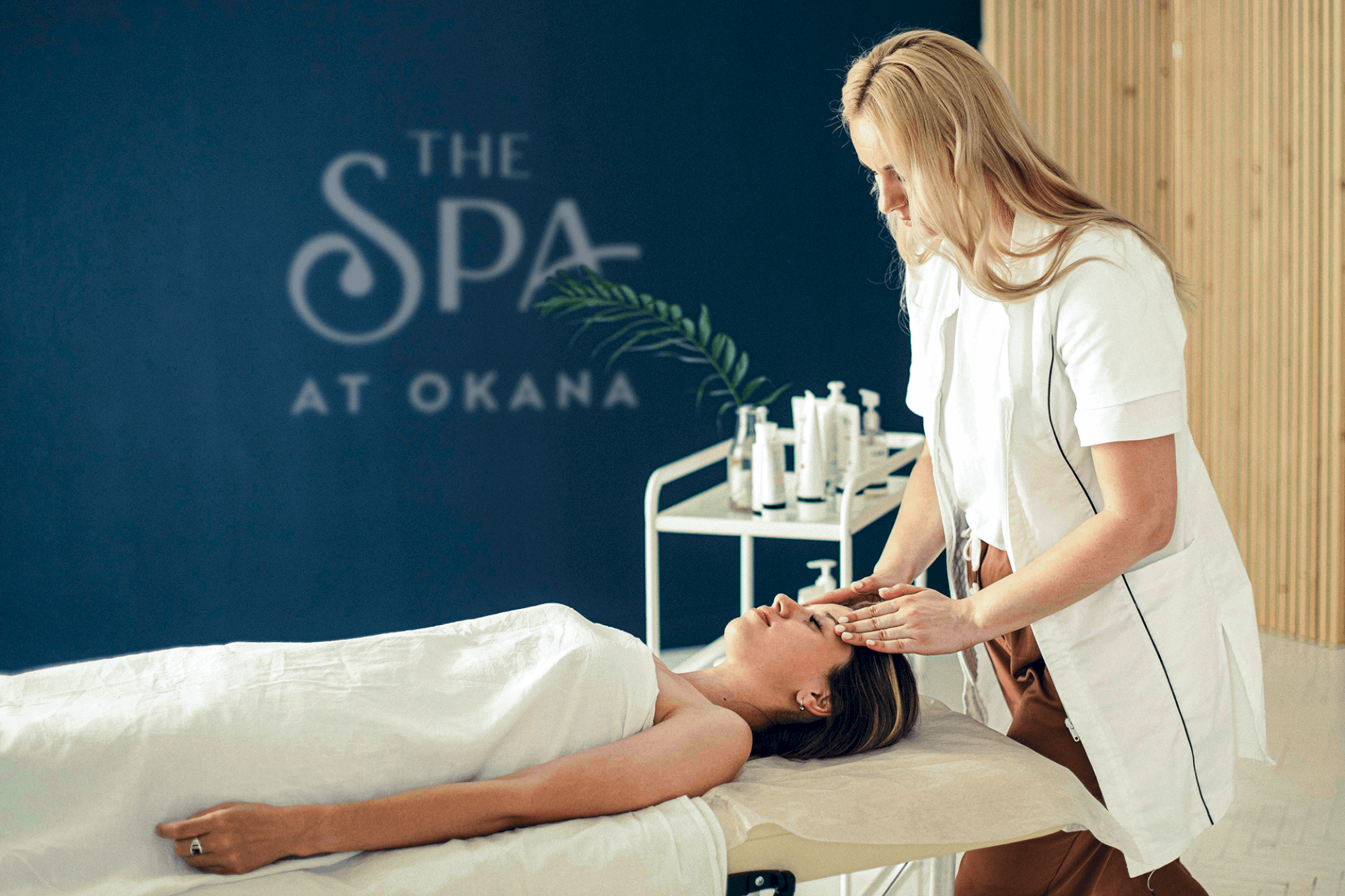 A spa therapist is massaging a client's face in a serene room with the text "The Spa at Okana" visible in the background.