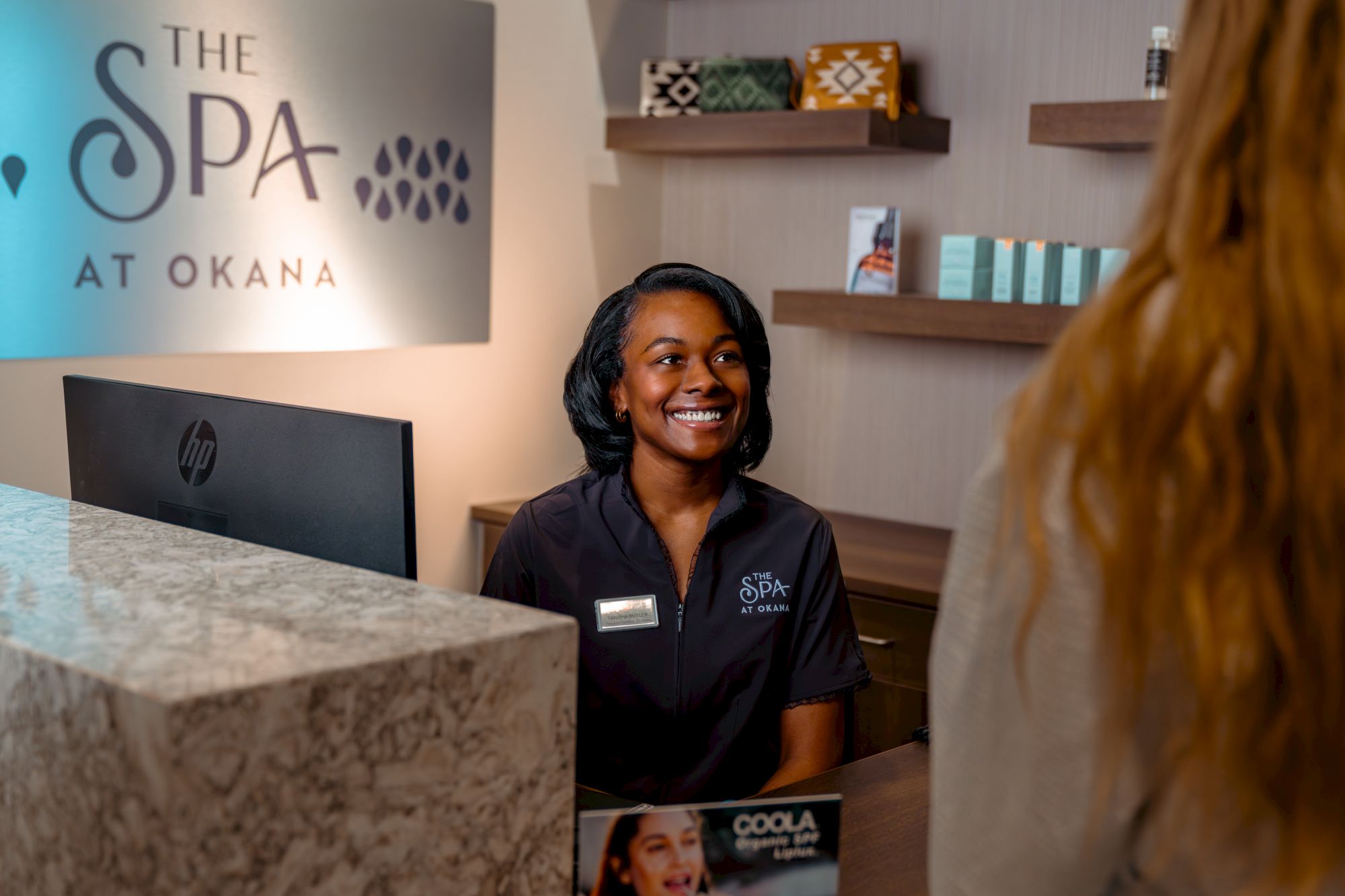 A smiling receptionist at "The Spa at Okana" desk interacts with a visitor. The setting is welcoming and professionally designed for spa services.