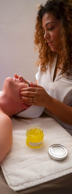 A person receives a facial massage from a therapist; a jar of yellow cream is nearby.