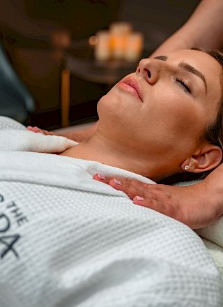 A person is receiving a neck massage while wearing a robe with a logo. Relaxing spa scene with candles in the background for ambiance.